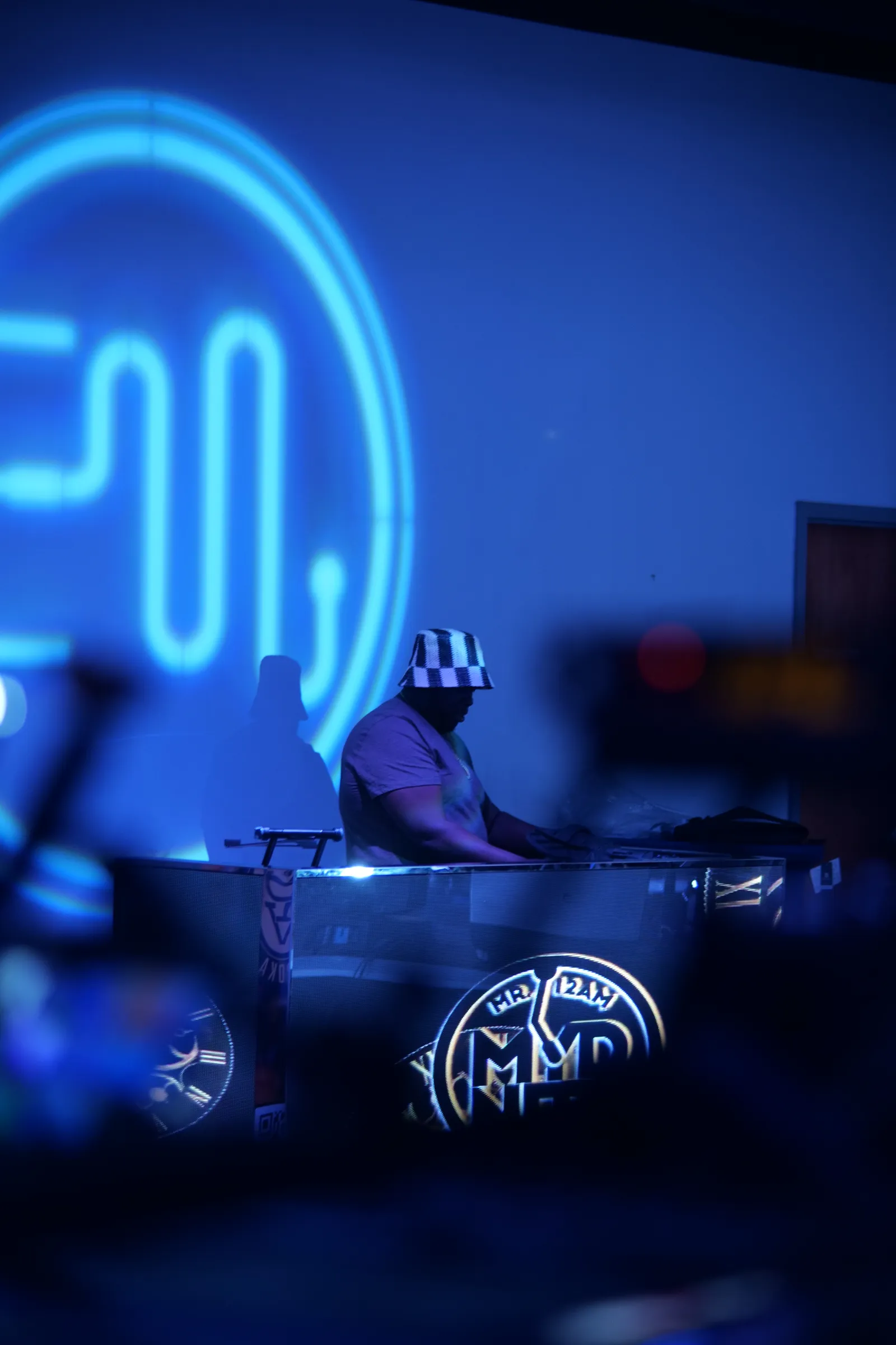 A DJ in a checkered bucket hat spins behind a lit booth at Elegance at Event Masterz, with the venue's blue neon EM logo glowing on the wall during a Charlotte live music night.